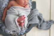 light gray swaddling wrap with baby's feet sticking out. blue wooden bowl, faux fur and gray layering fabric